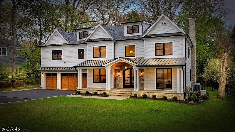 A 2025 center hall colonial at 15 School Avenue, photographed at dusk: white shake siding, warm timber garage doors, stone entry, every window glowing amber over a manicured front lawn.